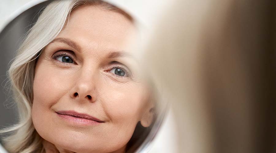 Femme mature souriante se regardant dans un miroir, illustrant l'effet raffermissant et lissant des soins visage anti-âge bio et naturels de IUM Paris.