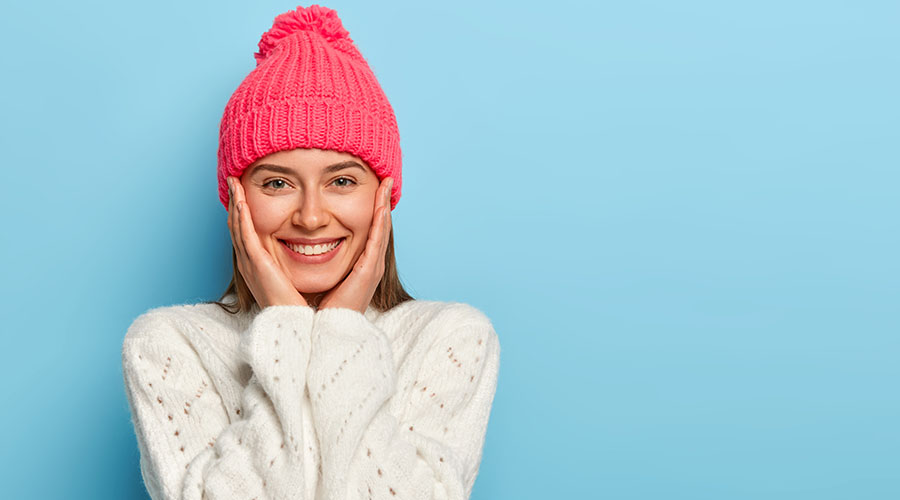 Femme souriante en bonnet rose et pull d'hiver, représentant une peau lumineuse et protégée grâce à une routine de soins naturels et bio adaptée à l'hiver. 
