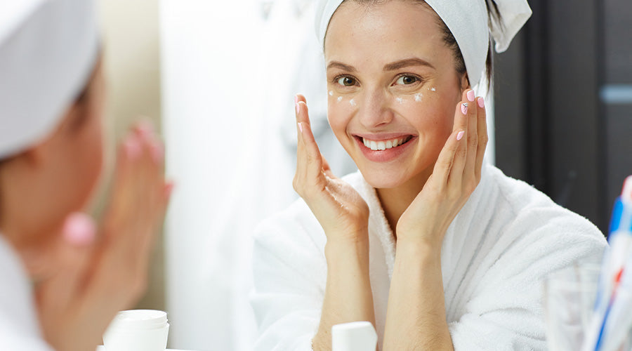 Femme applique de la crème ium bio sur le visage dans une sale de bain en se regardant dans un miroir