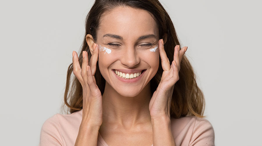 Femme souriante applique de la creme IUM sous les yeux avec ses doigts sur fond gris