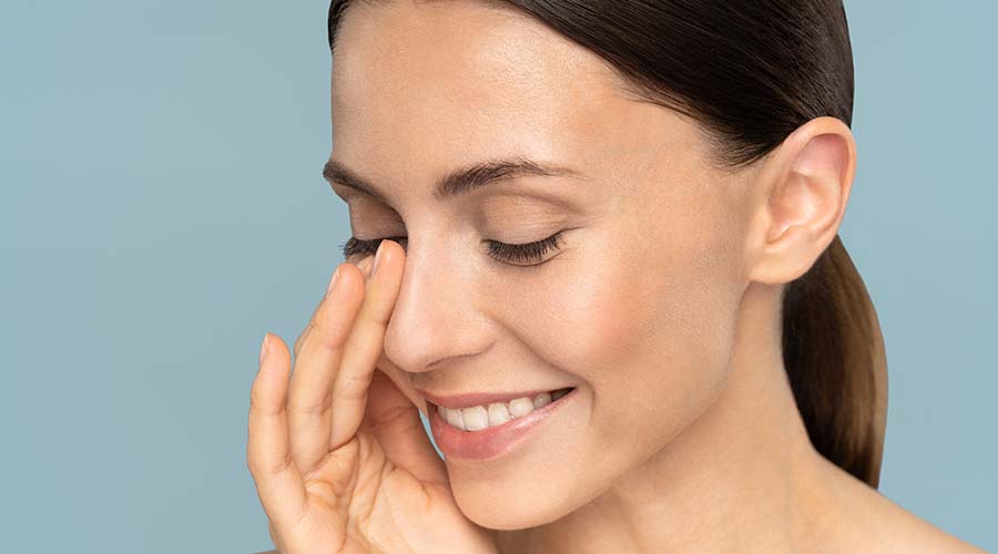 Visage femme brune quarantaine se touche le nez en souriant sur fond bleu