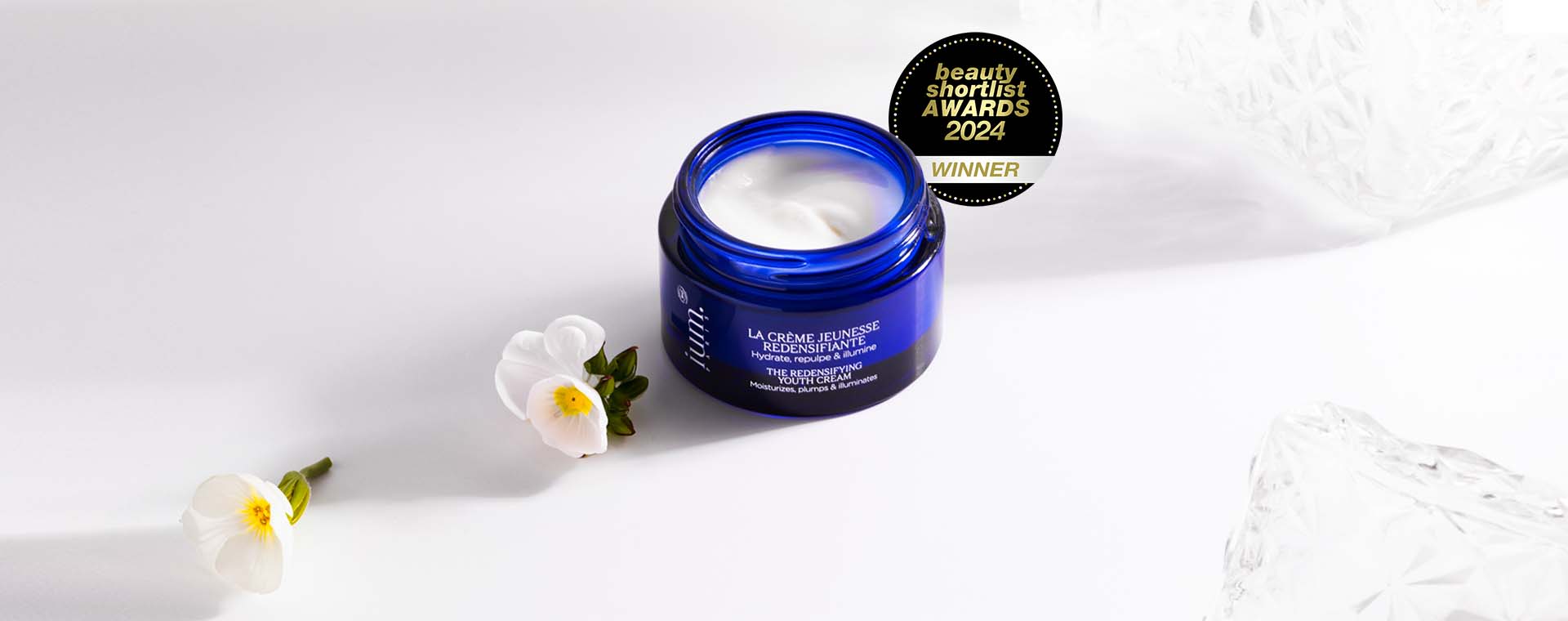 Pot ouvert de la Crème Jeunesse Redensifiante IUM Paris, entouré de fleurs blanches, lauréate du Beauty Shortlist Awards 2024, soin visage bio naturel, hydratant et repulpant.