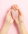 Mains de femme vues de haut qui tiennent une pierre de massage gua sha en quartz rose ium sur fond rose